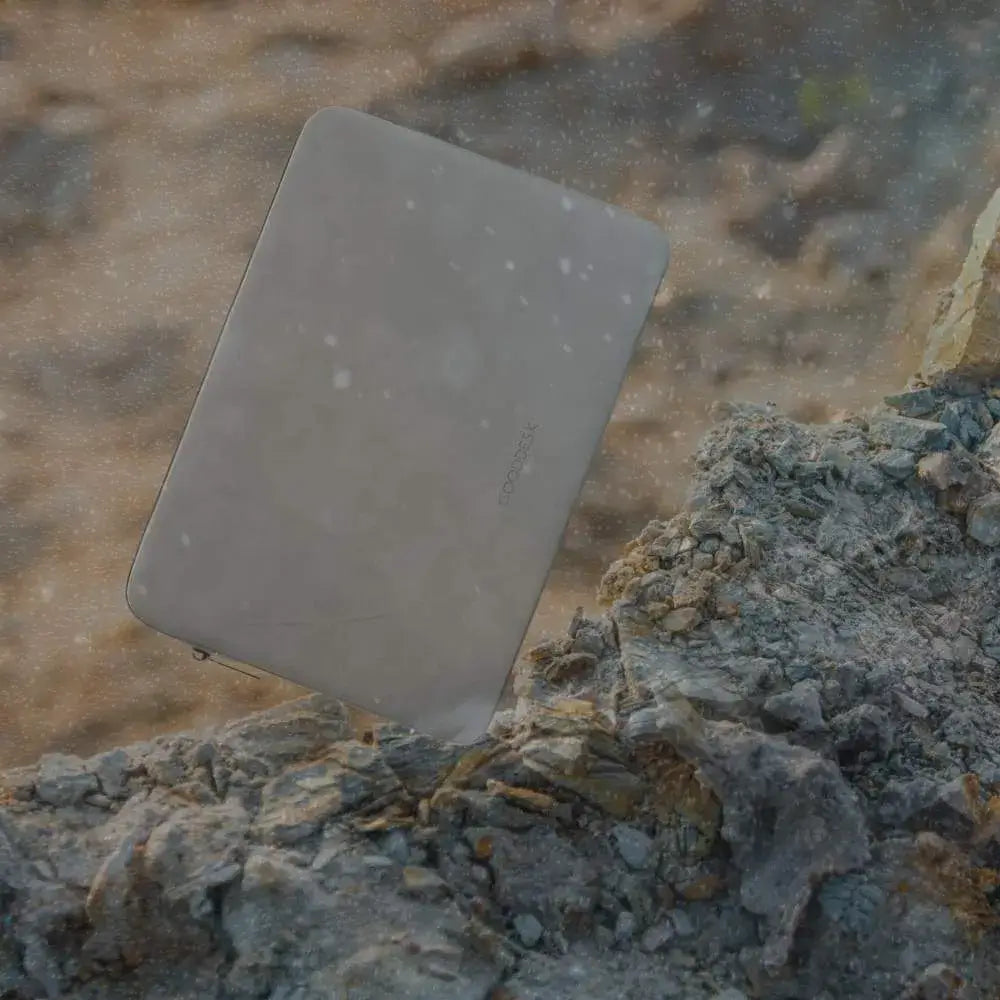 Eine graue Laptop-Hülle mit dem Logo 'GOODDESK' lehnt auf einem felsigen Untergrund, während im Hintergrund feine Schneeflocken oder Staubpartikel in der Luft schweben.