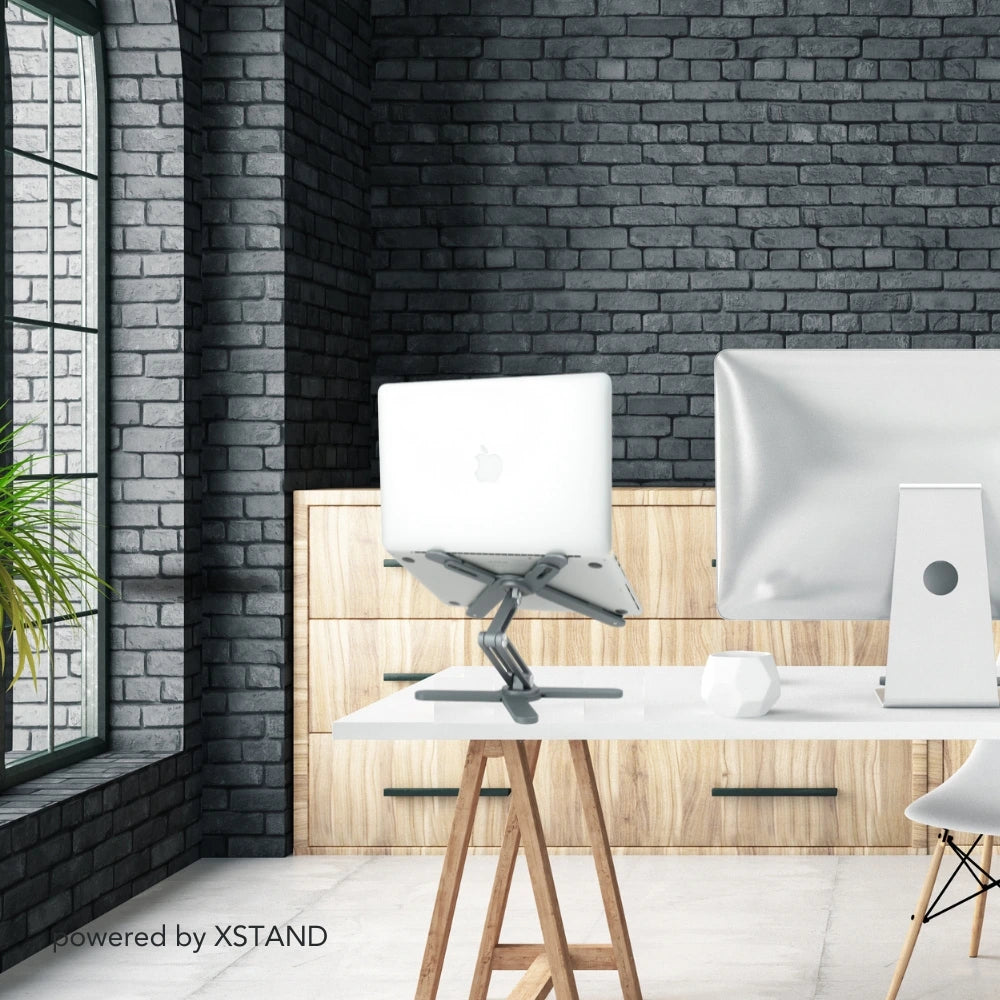 Moderner Arbeitsplatz mit XSTAND Laptophalter und einem MacBook, präsentiert in einem stilvollen, industriellen Bürodesign mit Backsteinwand und Holzelementen.